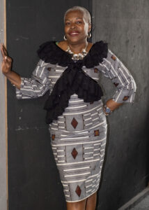 A black woman with short grey hair, black scarf, grey patterned dress, standing in front of a black wall
