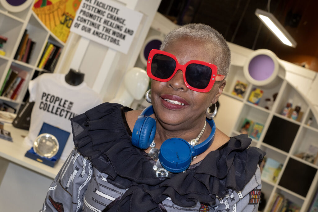 A black woman with short, grey hair, wearing red sunglasses, and blue headphones round her neck, black scarf.