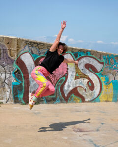A young white woman wearing black t-shirt and pink and yellow jogging bottoms leaping into the air, in front of a wall containing street art