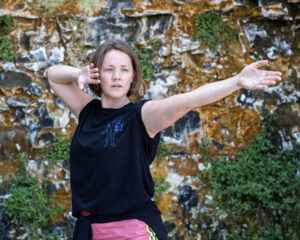 A young white woman wearing a black t-shirt and pink jogging bottoms, holds a dance pose, one arm outstretched, one arm bent, close to her head.