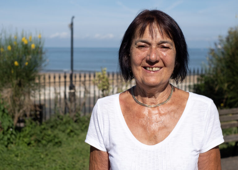Gabrielle Wilson is a white woman with short dark brown hair. She is wearing a white t-short and stands in front of a view of Margate seafront