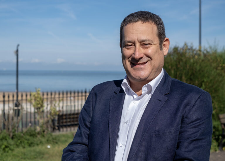 A white man with short hair wearing a white shirt and a navy blue suit jacket stands in front of a seaside backdrop