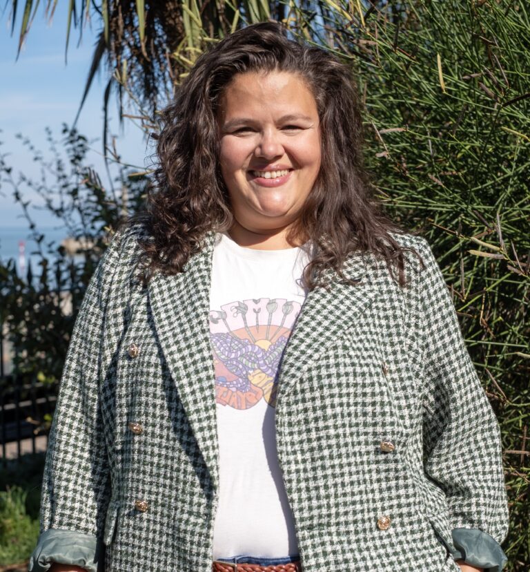 A young mixed race woman with long curly dark brown hair, wearing a checked jacket and a white t-shirt standing in front of some foliage