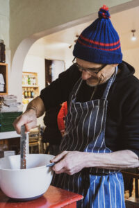 A white man wearing a blue hat with a red stripe, black top and blue and white striped apron, stirring a mixture in a white mixing bowl.