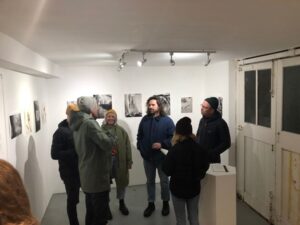 A group of people wearing outdoor jackets and hats gathered in a circle inside a small exhibition with pictures on a white wall